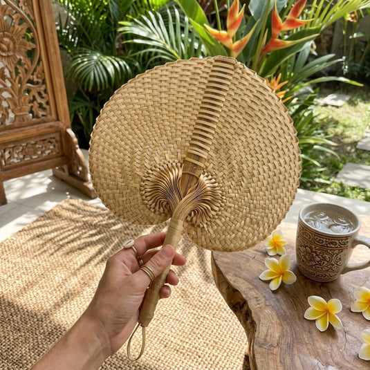 Handwoven Balinese palm fan held by hand in a tropical garden setting